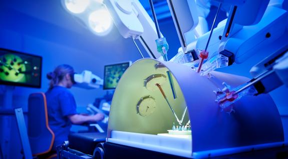 Im Vordergrund sieht mal Roboterarme, gesteuert von einer Chirurgin im Hintergrund, welche mit Hilfe von Trainingsmaterialien die Handhabung des OP-Roboters erlernt.