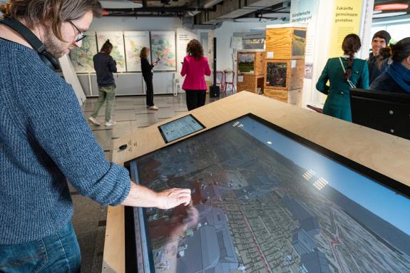 Ein Mann mit einem blauen Pullover und Brille bedient ein großes Touchscreen-Display, das die 4D-Browser Anwendung zeigt. Im Hintergrund sind mehrere Personen zu sehen, die sich eine Ausstellung ansehen. An den Wänden hängen Karten und Informationsplakate. Der 4D-Browser mit seinen historischen Stadtansichten wurde extra für den Touch-Screen optimiert und fügt sich dabei in eine Reihe ähnlicher Anwendungen ein, die das 3D-Stadtmodell als Grundlage für die Visualisierung und Interaktion nutzen.