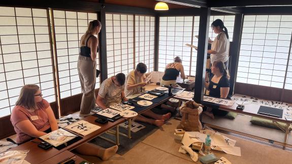 : Auf dem Foto sitzt eine Gruppe von 6 Studierenden auf Tatamimatten in einem traditionellen japanischen Daimyo-Haus und begutachtet Kalligraphien auf einem niedrigen Tisch, die sie davor angefertigt haben. Auf den Kalligraphien sind verschiedene japanische Kanji zu sehen.