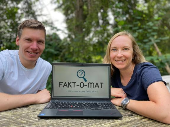 Zwei Menschen sitzen neben einem Laptop, auf dessen Bildschirm der Fakt-O-Mat zu sehen ist.