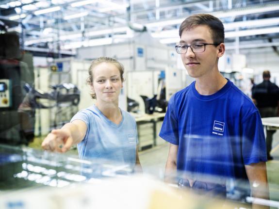 Ein Mädchen und ein Junge mit blauen Jenoptik-Shirts schauen konzentriert auf etwas in der Fertigungsumgebung, sie zeigt mit dem Zeigefinger auf etwas außerhalb des Bildes